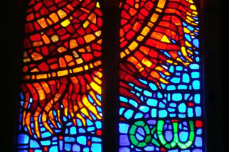 Stained glass window depicting a red/orange/yellow sun against a blue sky with Greek letters Alpha and Omega