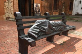 Jesus depicted sleeping on a park bench, covered in a blanket or robe. Identified as Jesus only by crucifixion wounds in his bare feet.