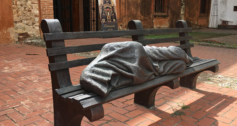 Jesus depicted sleeping on a park bench, covered in a blanket or robe. Identified as Jesus only by crucifixion wounds in his bare feet.