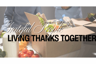 Grateful People: Living Thanks Together. A box of food donations sites on a table with two volunteer s in the background.