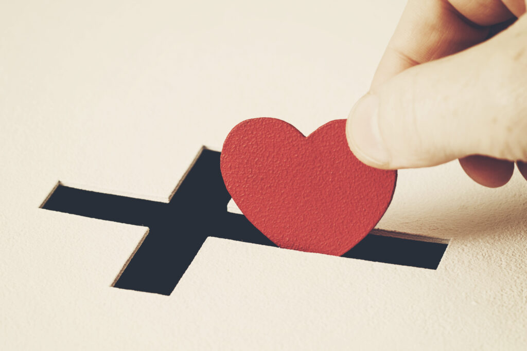 Hand placing a cutout heart into a cross-shaped hole, reminiscent of placing a coin into a child's piggy bank.